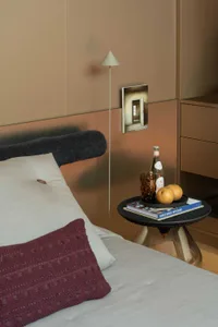 Modern bedroom corner featuring a bed with white and burgundy pillows, a black textured side table with brass base, a minimalist white floor lamp, and warm metallic wall panels.