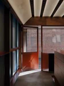 Modern minimalist entryway featuring dark walnut wood paneling on walls and ceiling, a terracotta-colored geometric pivot door standing open, and a view of a brick wall outside.