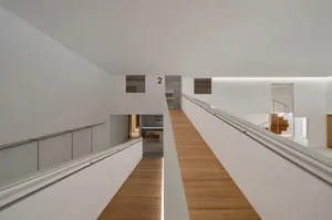 Looking down a modern white hallway with light oak flooring, featuring a floating staircase with wooden treads and metal railing, leading to lower levels with recessed lighting.