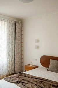Child's bedroom corner showing a wooden bed with textured pillow, brown throw blanket, oak nightstand with white sculpture, and playful giraffe and geometric patterned curtains allowing natural light.