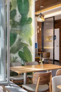 Modern restaurant dining area featuring a large tropical leaf mural on a ribbed glass partition, light wood tables, and mixed wood and upholstered chairs under pendant lighting.
