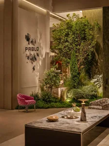 Modern lobby lounge featuring a large indoor garden with lush trees and plants, a marble coffee table, a vibrant pink patterned armchair, and a geometric patterned sofa under warm ambient lighting.