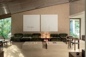 Modern living room featuring two forest green velvet modular sofas, beige stone coffee tables, warm wood panel walls, large abstract white artwork with the word PAUSE, and views of lush greenery outside.