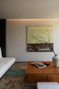 Modern living room featuring a sculptural wavy wooden coffee table, large abstract landscape painting in green and earth tones, patterned Persian-style rug, and a textured ceramic vase with flowering branches.
