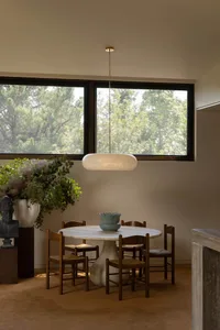 Modern dining room featuring a round travertine table, four dark wood woven-seat chairs, a large alabaster pendant lamp, and a large window overlooking green trees.