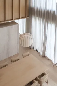 Modern minimalist dining room featuring a long light oak table, beige leather chairs, a large spherical paper lantern pendant, light wood cabinetry, and sheer white curtains.