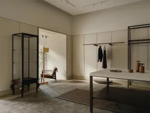 Modern minimalist entryway featuring a tall dark walnut glass vitrine, a limestone-top console table, open wardrobe system, and a vintage armchair visible in the adjacent sunlit room.