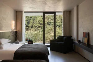Modern minimalist bedroom featuring a large bed with white linens and dark throw, black armchair, textured plaster walls, and expansive glass doors opening to a dense forest view.