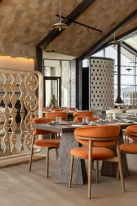 Modern restaurant interior with burnt orange velvet dining chairs, dark wood tables, geometric wooden room dividers, and a striking wood-paneled ceiling with pendant lights.