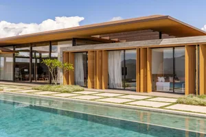 Modern luxury villa with extensive teak wood cladding, large glass windows, and an infinity swimming pool overlooking mountains under a blue sky.