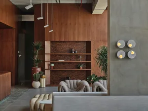 Modern lobby interior featuring a large walnut burl wood feature wall, built-in shelving, two curved bouclé armchairs, concrete wall sconces, and multiple potted plants.