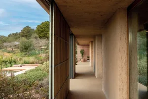 Covered outdoor corridor with rammed earth walls, wooden panels, and large glass doors overlooking a Mediterranean garden with pool and cacti.
