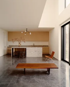 Modern minimalist open-plan kitchen and living area featuring polished concrete floors, a terrazzo kitchen island, light oak cabinetry, a live-edge oak bench, and a sculptural black chandelier.