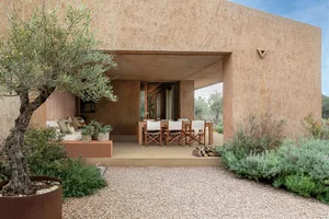 Modern Mediterranean courtyard patio featuring terracotta plaster walls, an olive tree, built-in outdoor sofa, wooden dining set, and gravel garden under a clear blue sky.
