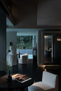 Modern minimalist living room featuring dark concrete walls, black floor, white linen armchairs, sculptural white bust on pedestal, and large opening revealing a swimming pool surrounded by lush greenery.