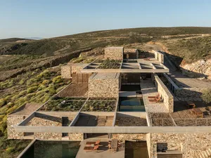 Aerial view of a contemporary stone and concrete courtyard villa with multiple terraces, infinity pools, and green roofs integrated into a rugged Mediterranean hillside.