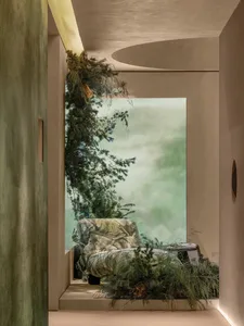 Modern biophilic interior featuring a stone-like armchair surrounded by lush evergreen plants, with soft natural light filtering through a large opening and curved plaster architecture.