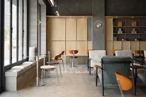 Modern cafe interior featuring concrete walls, light wood cabinetry, built-in window seating, round glass tables, and a mix of upholstered and molded wood chairs in green, orange, and neutral tones.