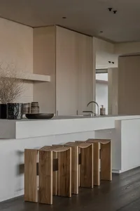 Modern minimalist kitchen interior with raw concrete island, curved walnut wooden stools, matte beige cabinetry, and dried floral arrangement.