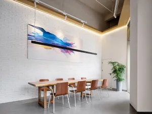 Modern dining room featuring a long live-edge wooden table, six walnut chairs, white painted brick wall, and a large colorful abstract painting illuminated by linear LED lighting.