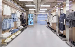 Long view down a modern clothing store aisle with stainless steel clothing racks, folded denim and white sneakers on display shelves, grid panel walls, and bright fluorescent ceiling lighting.