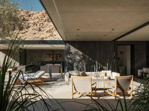 Modern desert villa outdoor lounge featuring concrete architecture, wooden lounge chairs, a light sofa, cacti, and a rocky mountain backdrop under bright sunlight.
