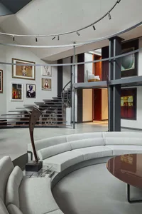 Modern double-height living room featuring a large curved light-grey sectional sofa, dark metal railing, floating staircase, and numerous framed artworks on white walls under a circular track lighting system.