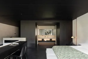 Modern dark-themed bedroom featuring a large bed with green patterned coverlet, black desk and chair in the foreground, and an open en-suite bathroom with double vanity visible in the background.