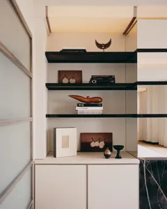 Modern minimalist built-in shelving unit with dark walnut floating shelves, white cabinetry, and carefully arranged books, wooden sculptures, and ceramic objects against a neutral wall.