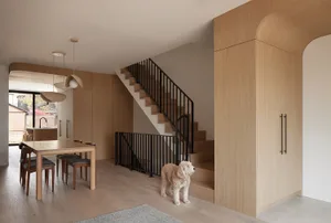 Modern dining room featuring a solid oak table and chairs, three pendant lights, light oak wall paneling, a black metal railing staircase, and a curly-haired dog standing on light wood flooring.