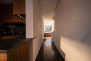View from a modern walnut kitchen counter looking down a narrow hallway with dark tiled floor toward a bright window at the far end.