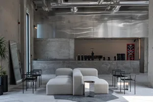 Modern industrial cafe lounge with raw concrete walls, a large rippled stainless steel feature wall, light grey modular sofas, black metal chairs, and exposed ceiling ducts.