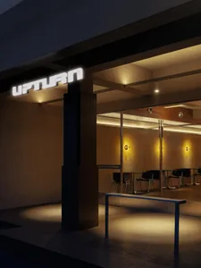 Night view of a modern cafe entrance featuring a large illuminated 'UPTURN' sign, a dark mosaic-tiled structural pillar, and a minimalist blue bench in the foreground.
