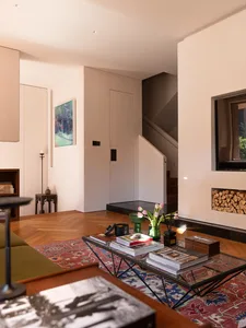 Modern minimalist living room with herringbone oak floor, colorful Persian rug, glass coffee table, built-in fireplace niche filled with firewood, and staircase in background.