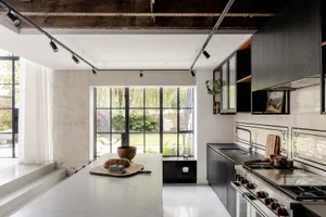 Modern industrial-style kitchen featuring a large concrete island, dark cabinetry, stainless steel range, and large black-framed windows overlooking a garden.