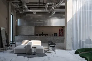 Spacious industrial-style cafe lounge with polished concrete floors, exposed ductwork, a large reflective stainless steel feature wall, white modular boucle sofas, black metal chairs, and a curved green velvet sofa in the foreground.