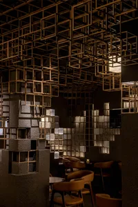 Interior of a modern restaurant featuring a dramatic ceiling installation of interlocking brass cuboid frames, textured concrete pillars with integrated lighting, and warm brown leather dining chairs.