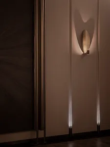 Modern luxury entryway featuring a textured brushed-brass leaf-shaped wall sconce casting dramatic light patterns on a neutral wall with dark wood paneling and marble floor.
