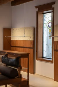 Walnut wood hall tree with metal hanging rod and textured panel, positioned next to a tall wooden-framed stained glass window featuring Art Nouveau patterns in purple, green and yellow.