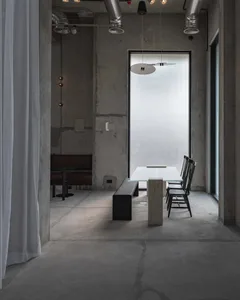 Industrial-style dining area with raw concrete walls and floor, a glass dining table on a concrete pedestal, black wooden chairs, a long black bench, and large frosted window allowing diffused natural light.