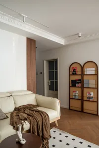 Modern minimalist living room featuring a cream leather sofa with a brown throw blanket, a round walnut coffee table with a white sculpture, and two arched walnut and rattan bookshelves against a white wall with crown molding.