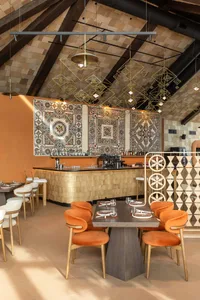Modern restaurant interior with orange velvet dining chairs, dark wood tables, a curved bar with golden tile cladding, large colorful patterned tiles on the wall, and geometric brass pendant lights hanging from a high beamed ceiling.