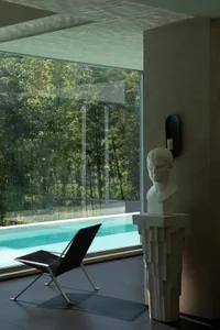 Modern minimalist indoor pool lounge featuring a black sling chair, a classical marble bust on a geometric concrete pedestal, large glass walls overlooking greenery, and dramatic natural light.