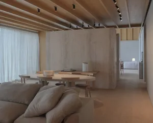 Modern minimalist dining room featuring a large curved light-wood table, matching wooden chairs, a prominent raw concrete partition wall, exposed wood beam ceiling with track lighting, and a soft beige sofa in the foreground.