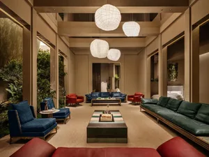 Modern luxury hotel lobby with deep blue, emerald green, and red velvet seating, layered coffee tables, and large spherical woven pendant lights in a double-height atrium with indoor plants.
