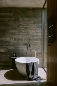 Modern minimalist bathroom featuring a white freestanding bathtub, dark concrete plank walls, a black stone stool with bath products, and a wooden door with hanging towels.