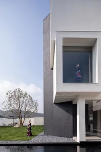 Modern minimalist concrete villa exterior with large square window framing a woman in colorful traditional indigenous clothing, a second woman in similar attire standing on the lawn beside a young tree, with a reflecting pool in the foreground.