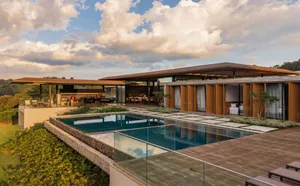 Modern hillside villa featuring an infinity pool, extensive teak wood decking, wooden louvered facades, and open-plan living areas overlooking rolling green hills under a dramatic cloudy sky.