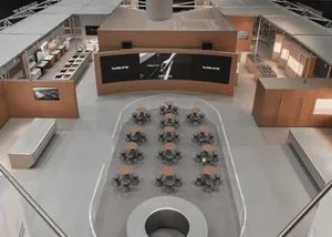High-angle view of a contemporary exhibition space with a large curved digital screen, oak paneling, and multiple round wooden tables with grey stools arranged in an oval formation on a light grey floor.