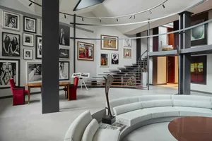 Modern double-height living room featuring a large curved white sectional sofa, dark structural columns, wooden accent furniture, and walls filled with framed black and white and color photography under a circular track lighting system.